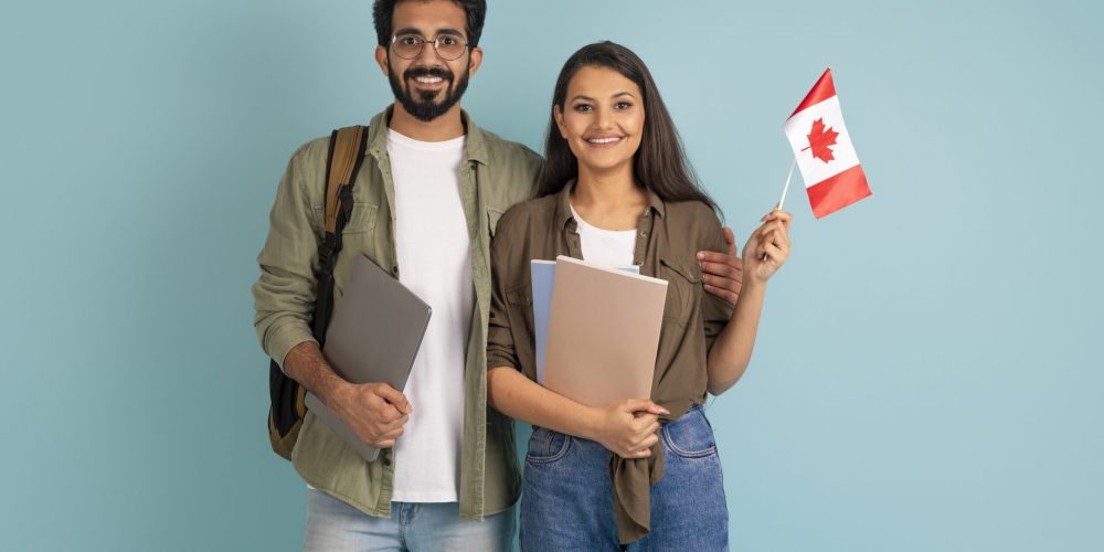 Education abroad, immigration to EU, language school. Cheerful multicultural young arab man and indian woman students with Canadian flag, books and laptop on blue background