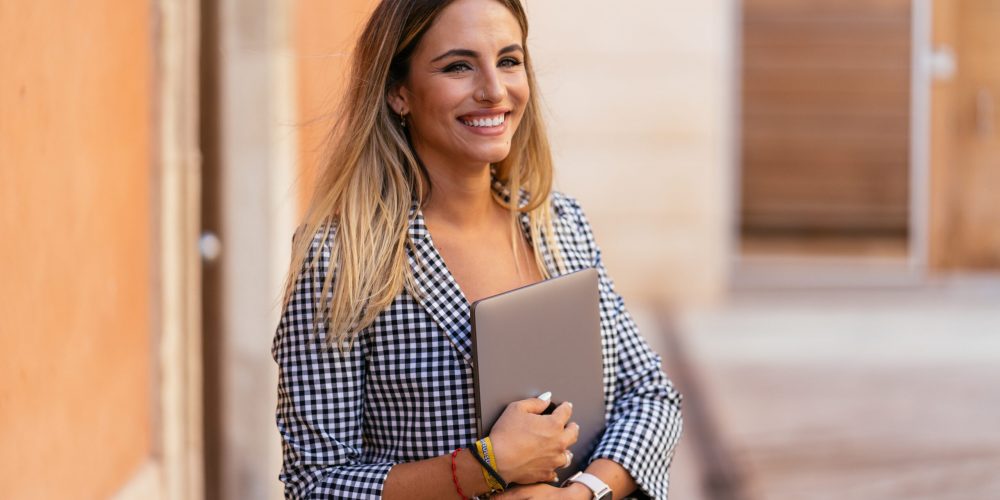 Glad female freelancer with netbook and in smart casual outfit standing in city street and looking at camera