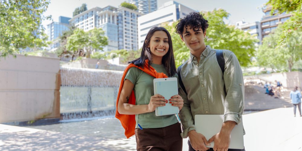 Two young college students are standing outside on a sunny day holding their laptop and tablet in a city park